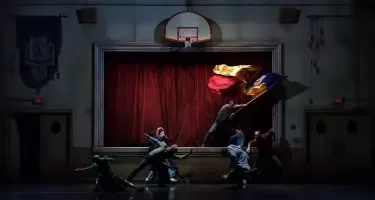 A group of six dancers in neutral coloured casual clothing strike scattered poses in front of a raised stage with a red curtain. Onstage one dancer waves a large red, yellow and blue coloured flag.