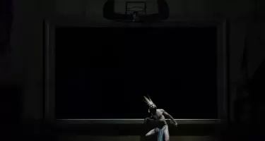 A solitary figure dressed in a long loin cloth and exaggeratedly tall crown leans against the edge of a raised stage in a dark room.