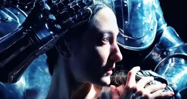 Renée Sigoiun in Kidd Pivot's "Assembly Hall". A close up on Renée's face, looking to the right. A person in a suit of medieval style armour stands close behind with one hand on Renée's head and one on her shoulder.