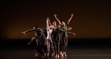 Group of dancers stands centre stage with arms raised. One dancer bends to the left with arms outstretched.