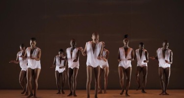 Seven Black women and Two Black menwith shaved heads and flowing white sleeveless tops stands in a group facing the camera, with arms raised slightly in front of them.
