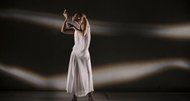 A Black woman with a shaved head in a flowing white dress stands alone onstage. Her upper body twists slightly to the right and her arms are raise in front of her face. There are waves of white light against black in the background.