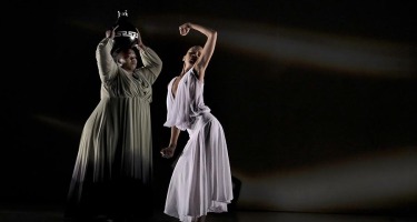 Two Black women stand together. One is in a flowing green dress and the other in a flowing white dress. The one in green holds a black vase on top of her head, the one in white stands beside her, bending and twisting slightly at the waist with one arm stretched below and one above. They stand against a black background with very faint wisps of white light.