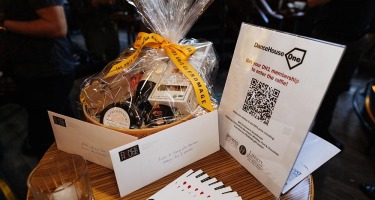 DanceHouse One social. A small, round, wooden table holds a gift basket full of cheese, a stand up sign with information about DanceHouse One, a glass of whiskey with an ice cube, and a fanned stack of white, black and red brochures.