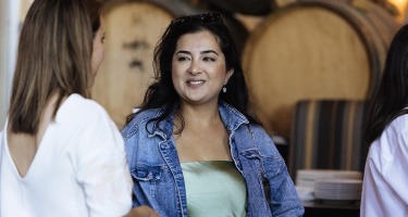 DanceHouse One social. An olive skinned woman with long, straight black hair, a green silk top and blue jean jacket talks to a light skinned woman with long, straight light brown hair and a white top. A stack of kegs sits in the background.