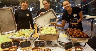DanceHouse One social. Three servers dressed in black show off dishes of food set up buffet style on a table.