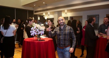 DanceHouse One social. A group of people stand around a room full of cocktail tables with  drinks. A man in a plaid shirt wih a dark beard raises a toast to the camera with a glass of wine.