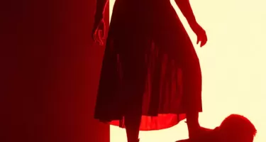 A woman in a loose, translucent dress stands on the back of a man who is crouched over. They are both backlit by yellow light and appear as red silouettes.