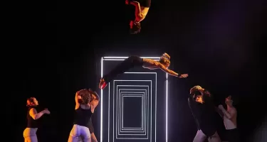 Gravity and Other Myths, The Mirror photo by Andy Phillipson. A group of dancers in tight white and black shorts and sleeveless black tops toss a female dancer in long black tights, a white sleeveless top and a blonde pony tail in the air. Another dancer in black tights and a sleeveless white top is suspended in the air mid somersault. A series of nested doorway-like rectangles made out of flourescent tubes is in the background.