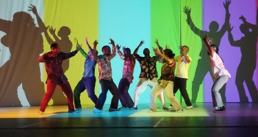 Via Katlehong Dance Company. Eight colourfully dressed dancers stand closely in a line with their arms raised. Behind them is a bright projected backdrop of wide bands of colour.
