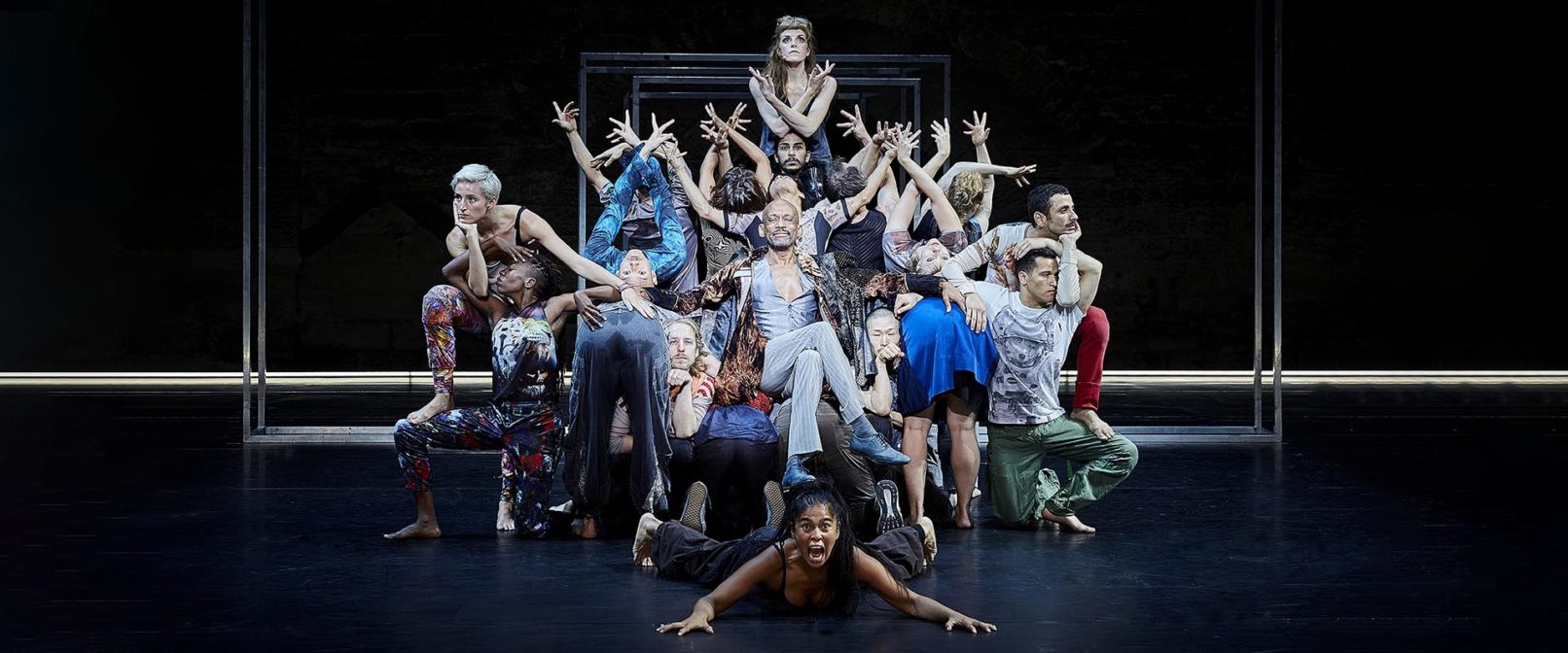 Babel. Group of people with arms raised closely surround man seated in chair.