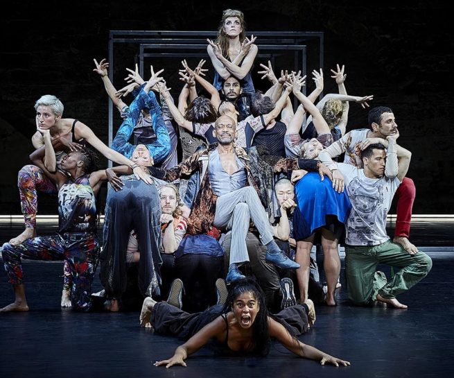 Babel. Group of people with arms raised closely surround man seated in chair.
