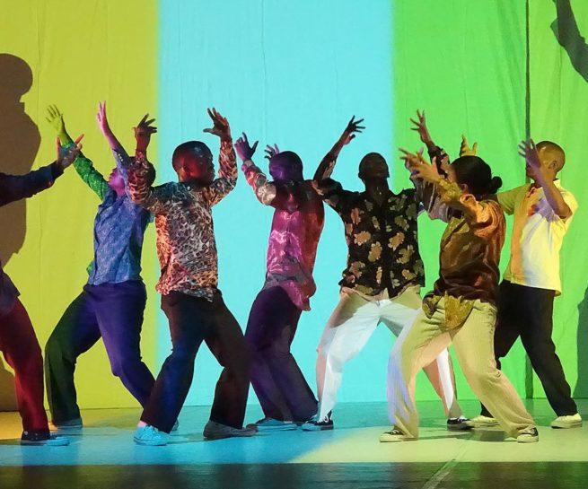 Via Katlehong Dance Company. Eight colourfully dressed dancers stand closely in a line with their arms raised. Behind them is a bright projected backdrop of wide bands of colour.