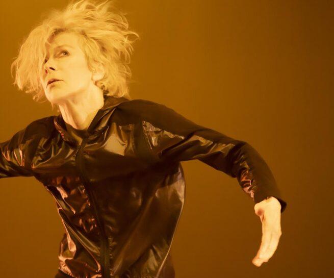 A woman with shaggy blond hair and a shiny black jacket faces the camera with her arms outstretched to her sides.