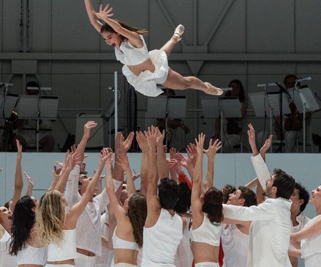 Paris Opera Ballet - Play. Large group of dancers dressed in white clothing stand in a group, a female dancer in white is thrown in the air by the group.