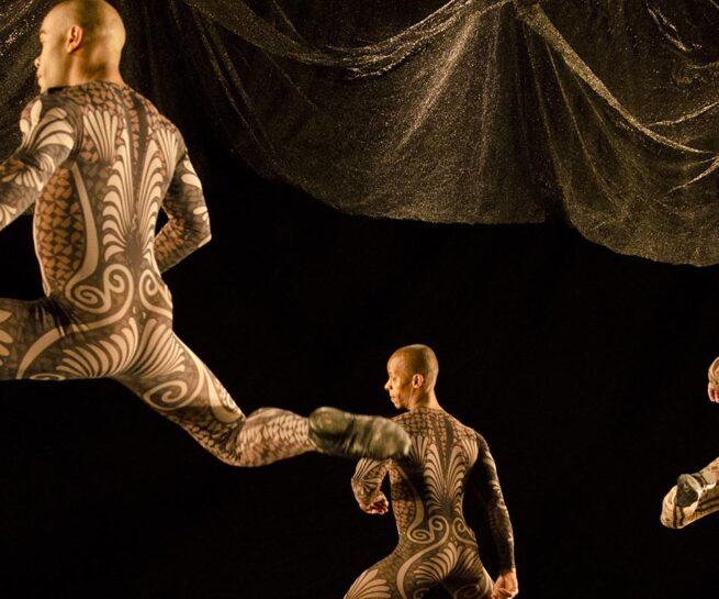 Grupo Corpo. Sem Mim. Three male dancers in skin tight leotards that resemble body tattoos leap on stage against a black backdrop and dark gold coloured curtain.