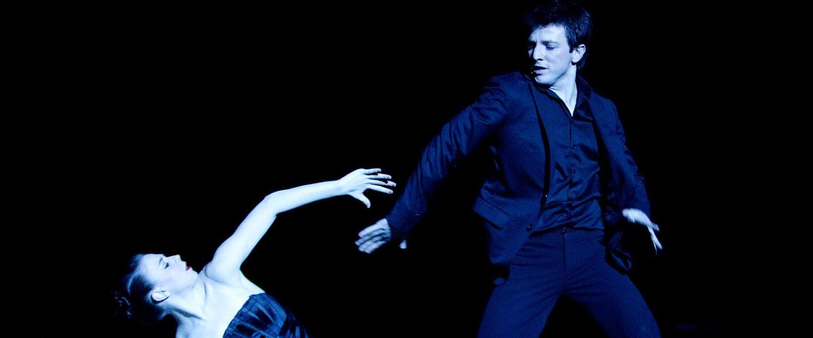 La la la Human Steps. woman in a black bustier reaches up to man in black pants, shirt and blazer.