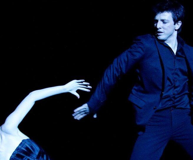 La la la Human Steps. woman in a black bustier reaches up to man in black pants, shirt and blazer.