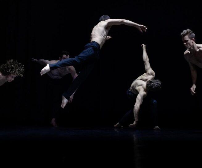 Circa. A group of five, shirtless, fair skinned male dancers in dark blue trousers. Three leap so that their bodies are parallel to the stage and the other two bend forward with arms stretched downward. They are lit against a dark background.
