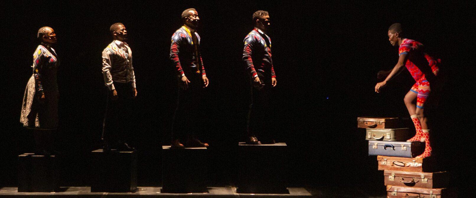 Gregory Maqoma and Thuthuka Sibisi. A Black man in red patterned socks, shorts and t shirt climbs steps made from stacked suitcases. Four other people in colourful clothing face him while standing on boxes in a row. The man on the suitcases is lit brightly, while the others are backlit. Everyone stands out against a black backgound.