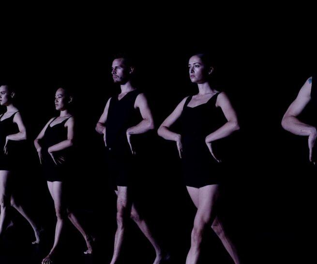 Batsheva Dance Company. Eleven fair skinned dancers in tight black sleeveless tops and shorts stand side by side in a line with their hands on their hips. They are facing slightly to the left of the frame. The dancer on the far right is closest to the camera with the others receding back into the frame to the left.