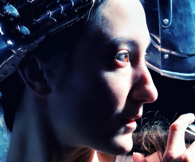 Renée Sigoiun in Kidd Pivot's "Assembly Hall". A close up on Renée's face, looking to the right. A person in a suit of medieval style armour stands close behind with one hand on Renée's head and one on her shoulder.
