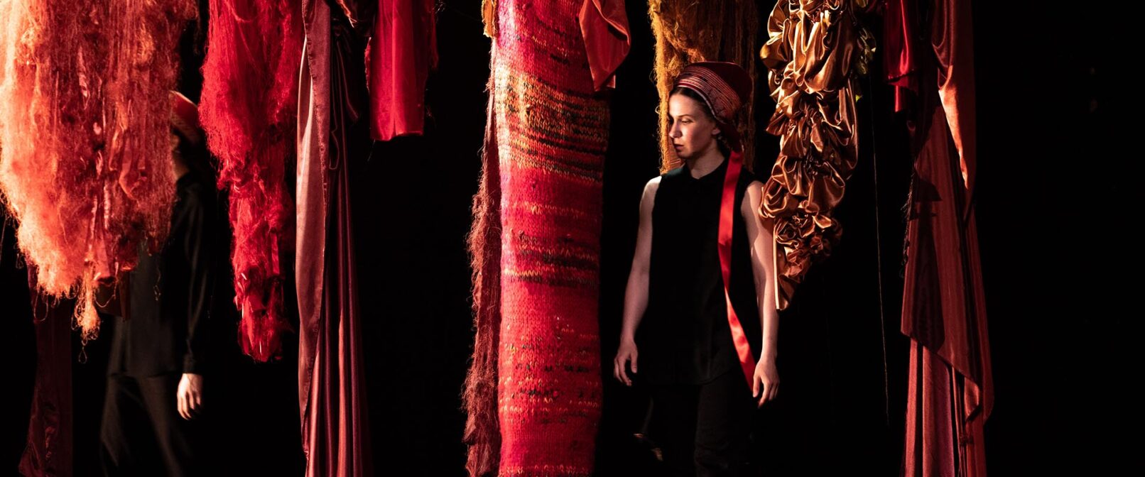 Elle Sofe Sara. A fair skinned woman dressed in a sleeveless black dress and red kerchief on her head stands amidst red patterned pieces of fabric that are hung from above. The effect is as if she is standing in a forest of fabric.