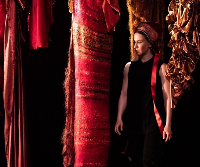 Elle Sofe Sara. A fair skinned woman dressed in a sleeveless black dress and red kerchief on her head stands amidst red patterned pieces of fabric that are hung from above. The effect is as if she is standing in a forest of fabric.