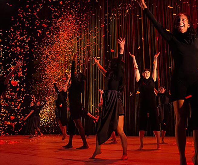 Nova Dance. A scene with a sense of joy that shows a group of dancers spread across a stage all wearing black. Some have their arms raised. The stage is lit red and the backdrop has splashes of red light against black.
