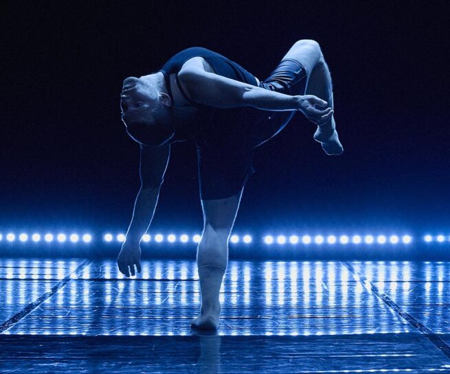 Guillaume Cote Danse. A dancer stands centre stage bending backwards with one leg raised and knee bent. The top half of the background appears dark, while the bottom half is a projection of blue light and shadow forming a row of narrow vertical bars.