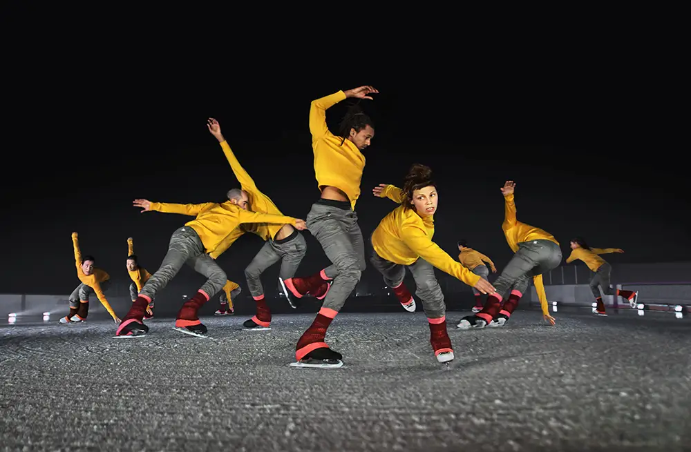Le Patin Libre. A group of mixed gender figure skaters dressed in grey pants, yellow long sleeve tops and pink leg warmers skate in a loose group, reaching with their arms in various directions.