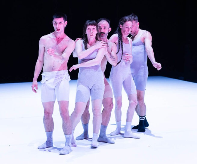 Compagnie Catherine Gaudet. Five fair skinned dancers stand grouped tightly together on a white stage, facing forward. They are wearing white and light grey tight sleeveless, and thigh length body suits.